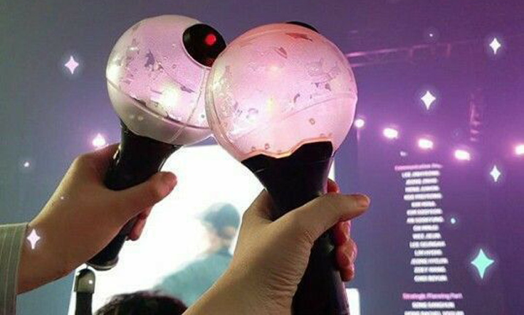 Dos lightsticks ARMY Bomb de BTS sostenidos en alto durante un concierto, con el escenario iluminado al fondo.